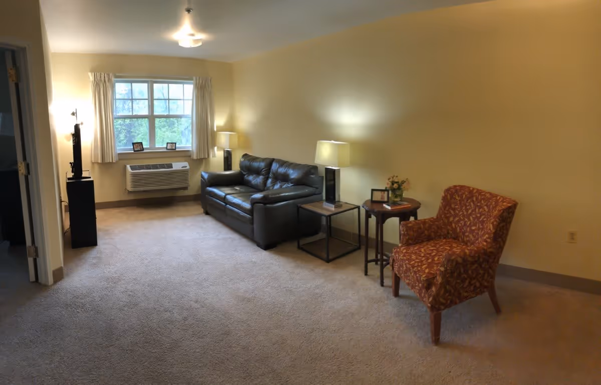 A living room with beige walls and carpet, featuring a black leather sofa, a patterned armchair, two side tables with lamps, a small round table with a flower vase and picture frame, a window with white curtains, and an air conditioning unit below the window.
