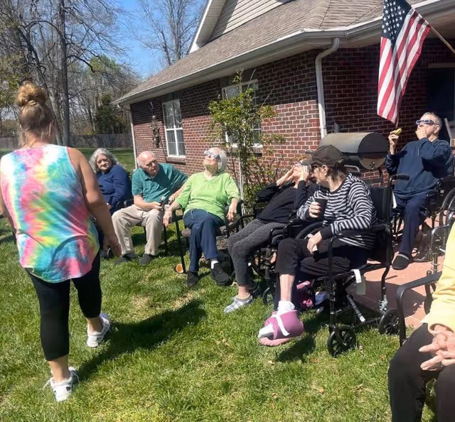 A group of elderly people sitting outside on chairs and wheelchairs on a grassy lawn next to a brick building. One person is standing and walking away from the group. An American flag is visible on the building. The weather is sunny with a clear blue sky.