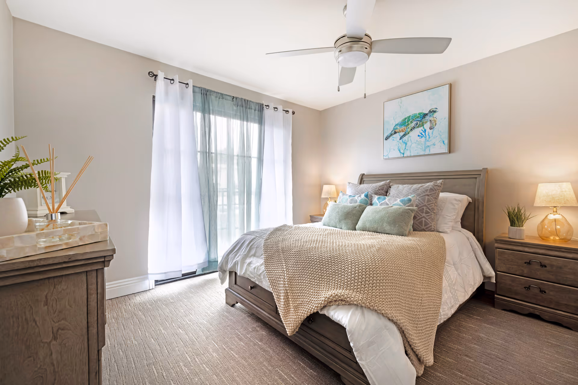 A well-lit bedroom with a large bed featuring multiple pillows and a beige knitted throw blanket. The room has light-colored walls, a ceiling fan, two wooden nightstands with lamps, a dresser with decorative items, and a window with sheer white and blue curtains. A framed picture of a sea turtle hangs above the bed.