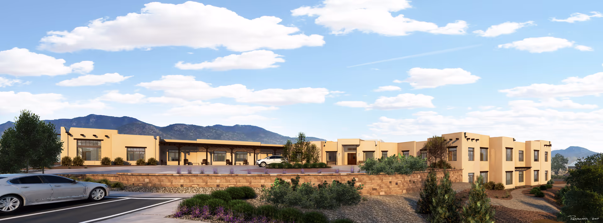 Exterior view of a single-story assisted living and memory care facility with a southwestern architectural style, featuring beige stucco walls, flat roofs, and large windows. The building is surrounded by landscaped greenery, desert plants, and a parking area with a silver car. Mountains and a partly cloudy sky are visible in the background.
