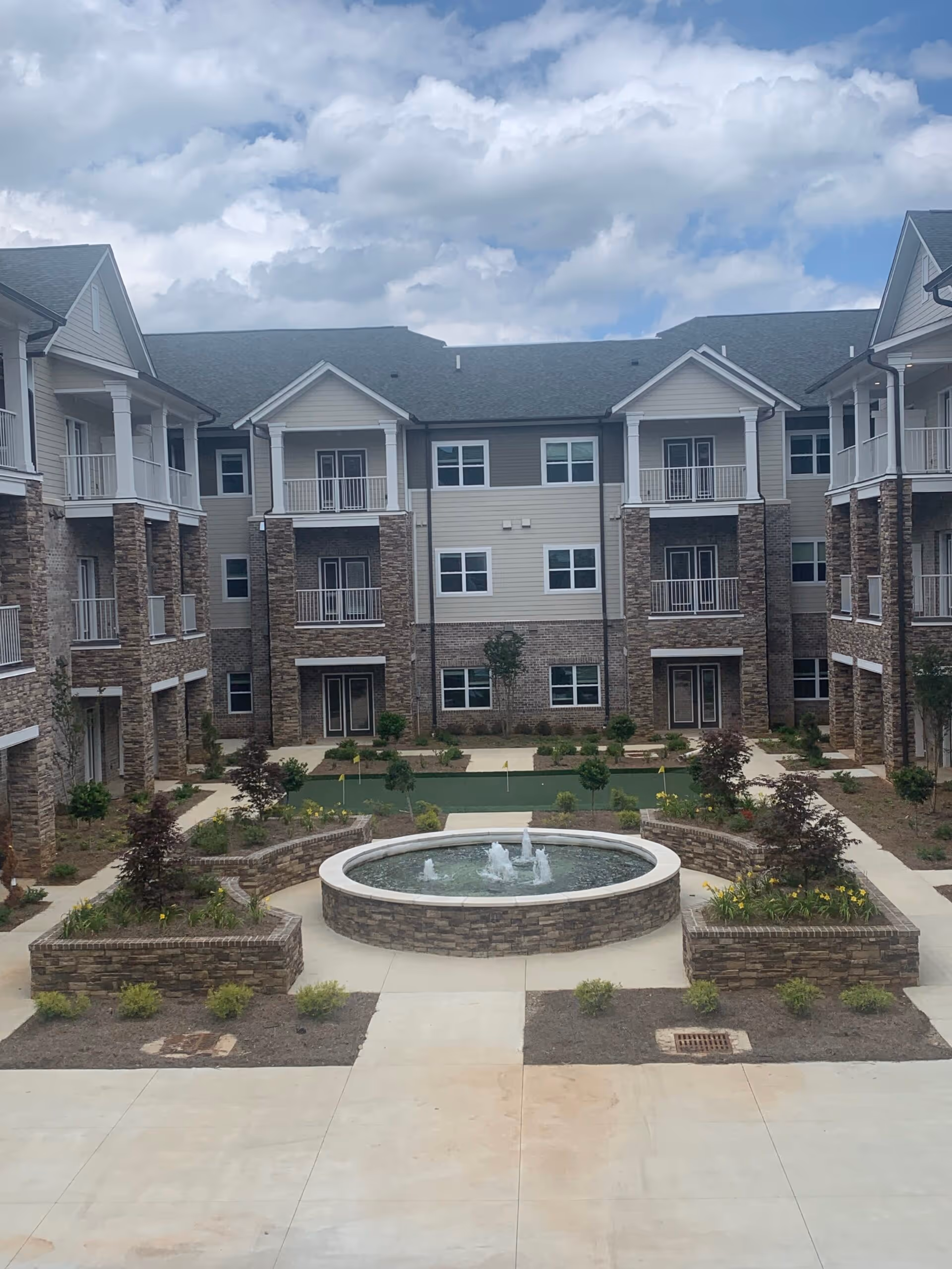 Outdoor courtyard area of a senior living facility with a circular stone water fountain in the center, surrounded by landscaped flower beds and small trees. The courtyard is enclosed by a three-story building with balconies and stone and siding exterior walls under a partly cloudy sky.