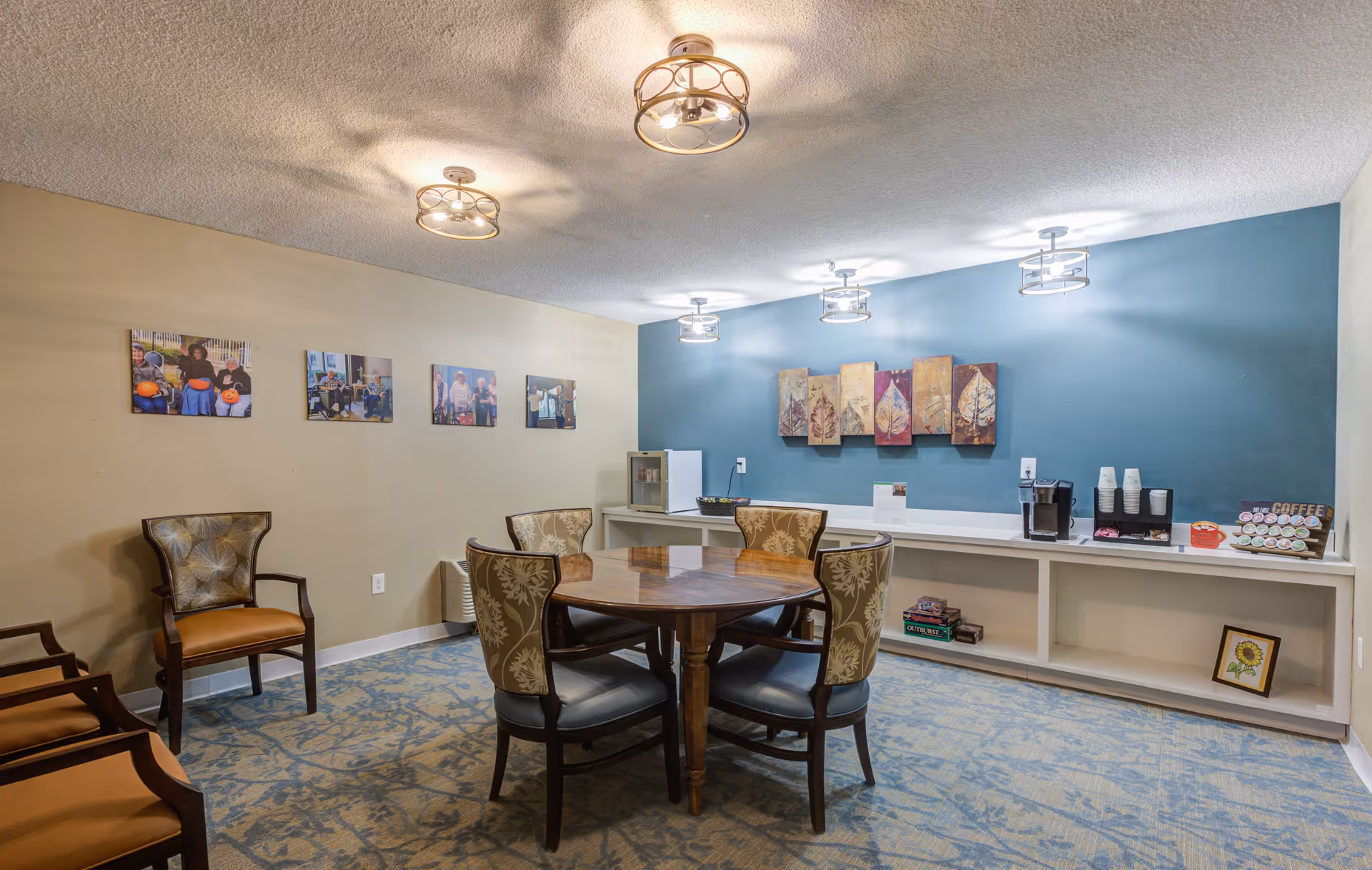 A cozy common area in a senior living community featuring a round wooden table surrounded by four upholstered chairs with floral patterns. The room has a blue accent wall with decorative leaf artwork and a counter with a coffee station, cups, and a small refrigerator. The opposite wall is beige with framed photos of people. The floor is carpeted with a blue and beige pattern, and the ceiling has modern light fixtures.