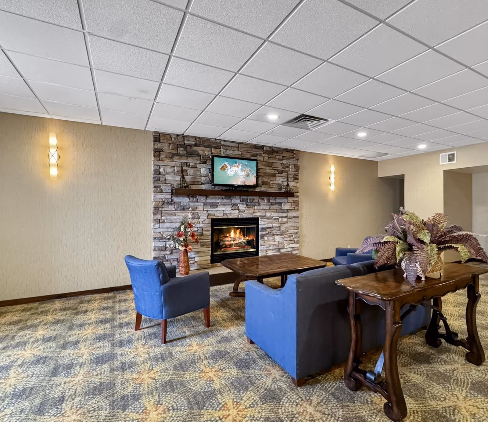 Cozy communal lounge with a stone fireplace and wall-mounted TV, blue chairs and sofas arranged around coffee tables.