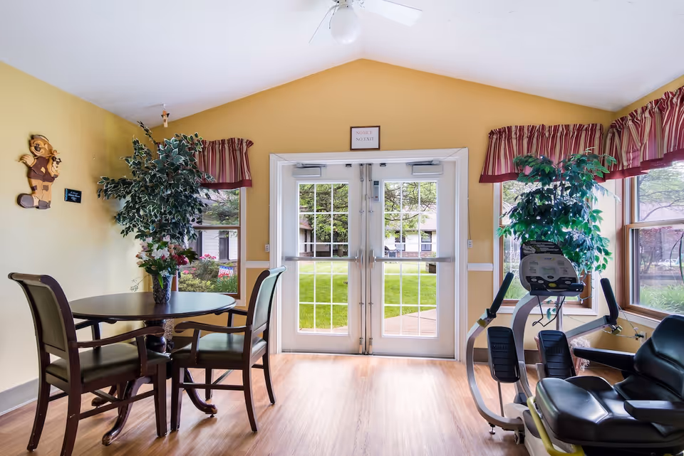 A bright room with yellow walls featuring a round table with four chairs and a vase of flowers on it, two large potted plants, an exercise machine, and double glass doors leading outside to a green lawn and trees. The room has wood flooring and windows with red and white striped valances.