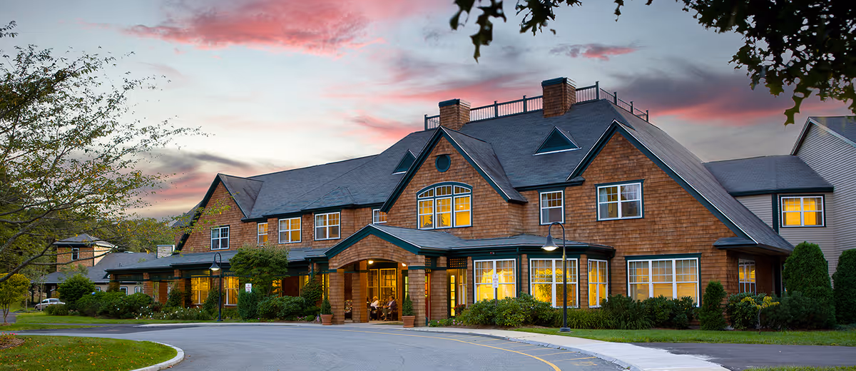 Exterior view of a large senior living facility building with warm lights glowing from the windows during sunset, surrounded by greenery and a curved driveway in front.