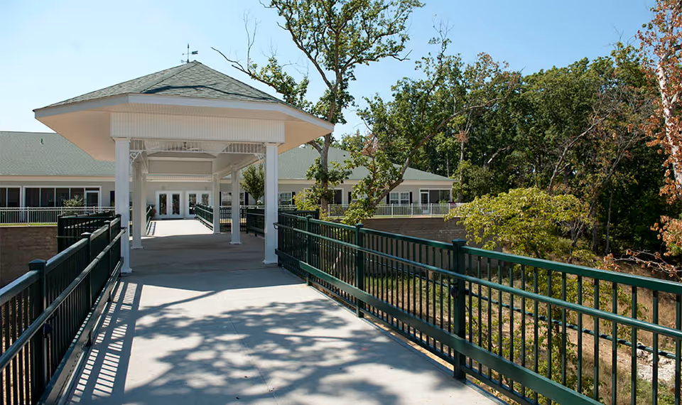 Covered walkway with green railings leading to the entrance of a single-story building set among trees.