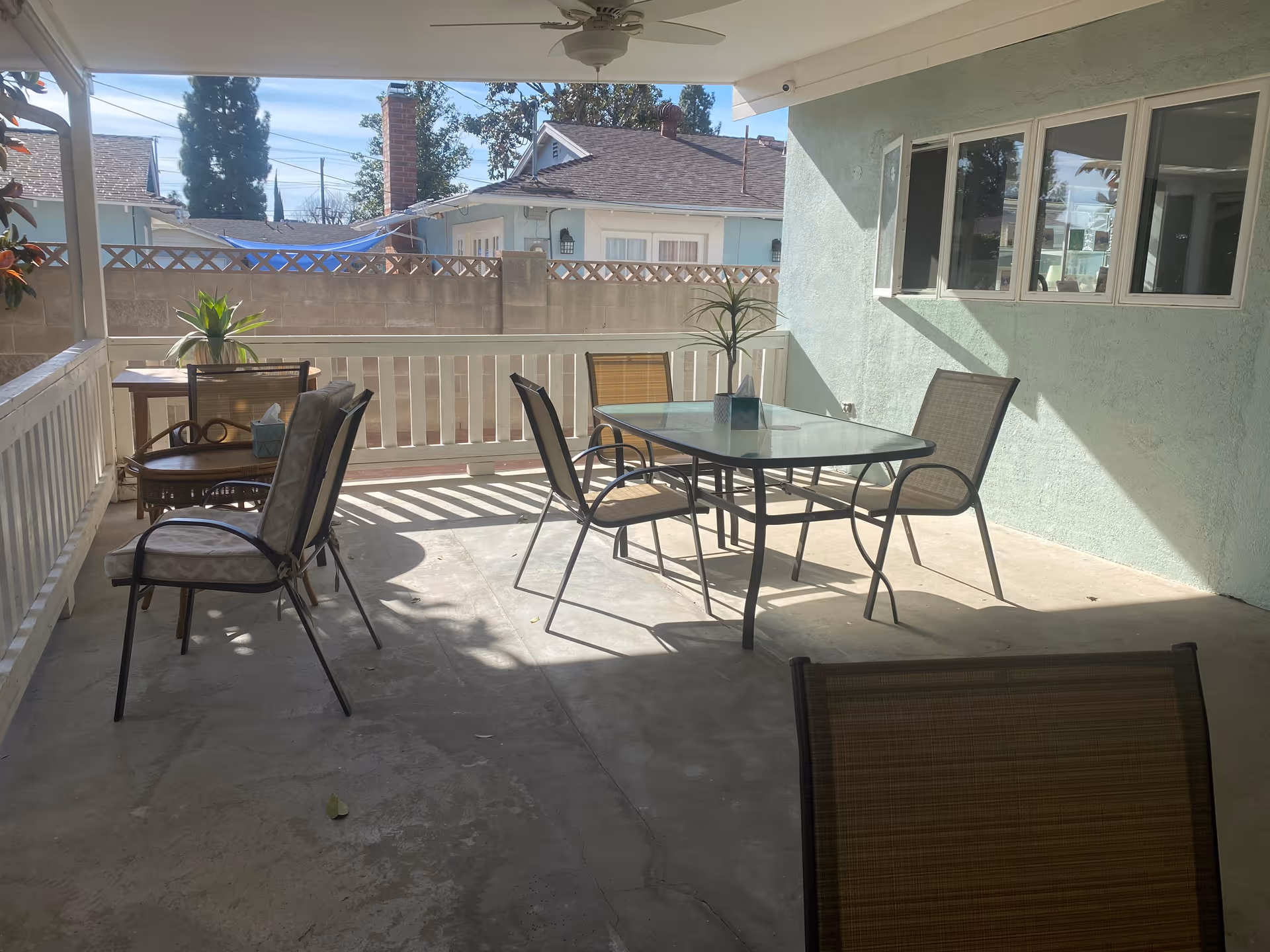 Covered outdoor patio area with a glass-top table and four chairs, additional seating with two cushioned chairs and a small table with a plant, surrounded by a white railing and a concrete floor, with neighboring houses and trees visible beyond a low brick wall.