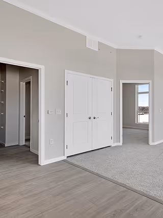 Interior view of a senior living facility showing a hallway with light gray walls, white double doors, and a carpeted room visible through an open doorway. The floor transitions from wood-style flooring in the hallway to carpet in the adjacent room. There is a window in the carpeted room letting in natural light.