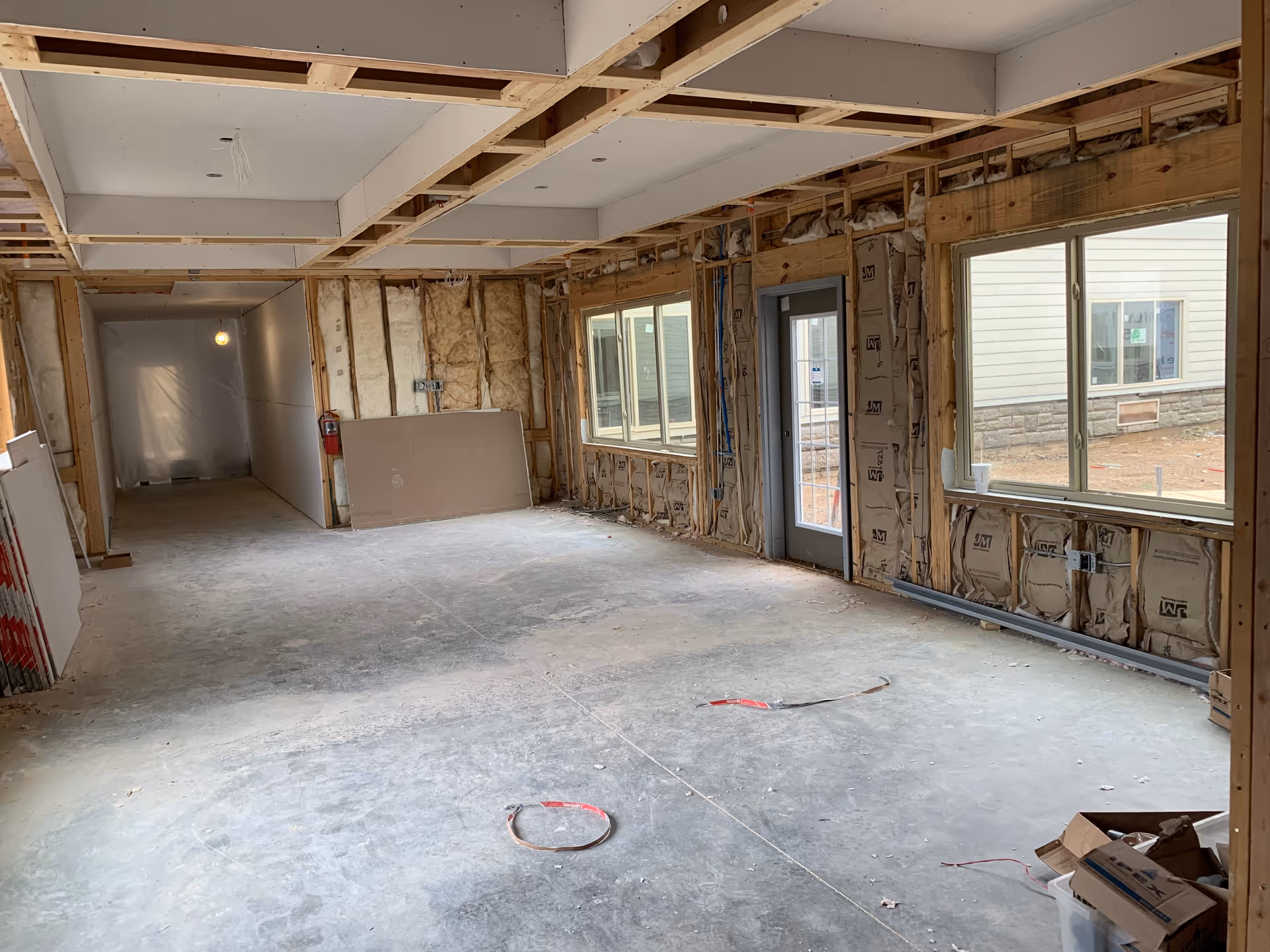 Interior view of a room under construction with exposed wooden framing, insulation, and drywall partially installed. The floor is unfinished concrete, and there are large windows and a door on the right side letting in natural light. Construction materials and tools are scattered on the floor.