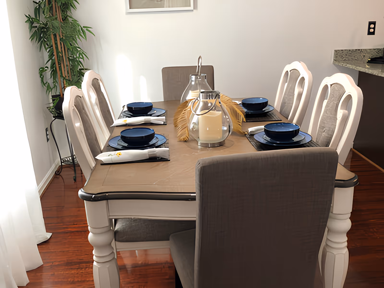 Dining room table set for six with blue dishes, candles and a decorative centerpiece and upholstered chairs on a hardwood floor.