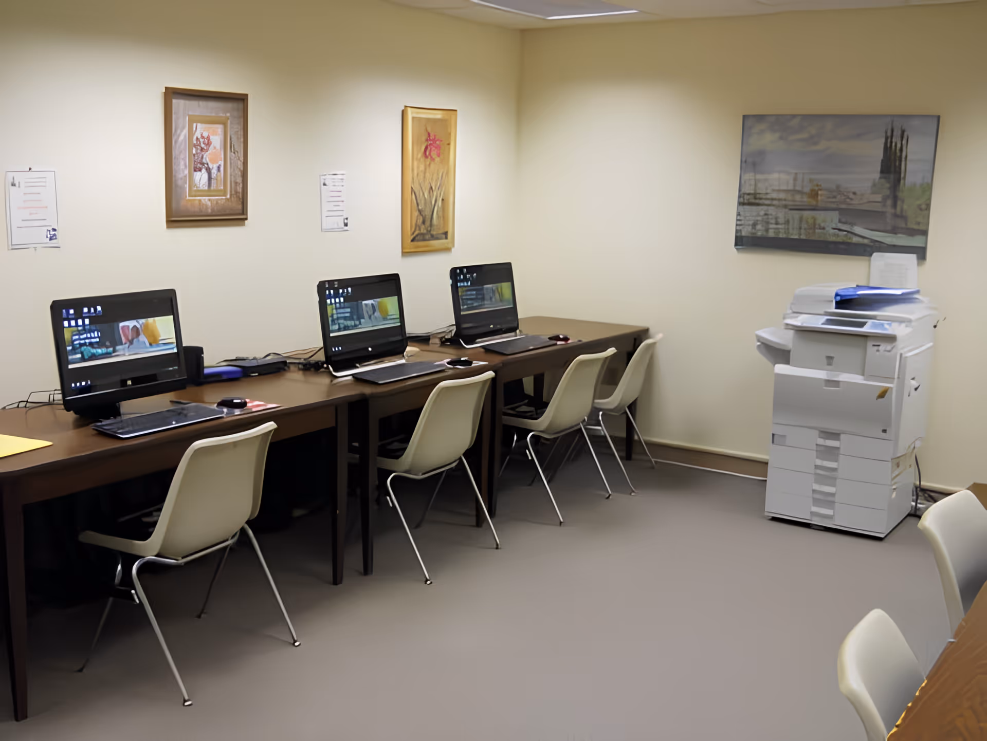 A small office or computer room with three desktop computers on a long brown table, each with a white chair. There is a large white printer or copier machine in the corner. The walls are beige with three framed artworks hanging. The floor is covered with light gray carpet.