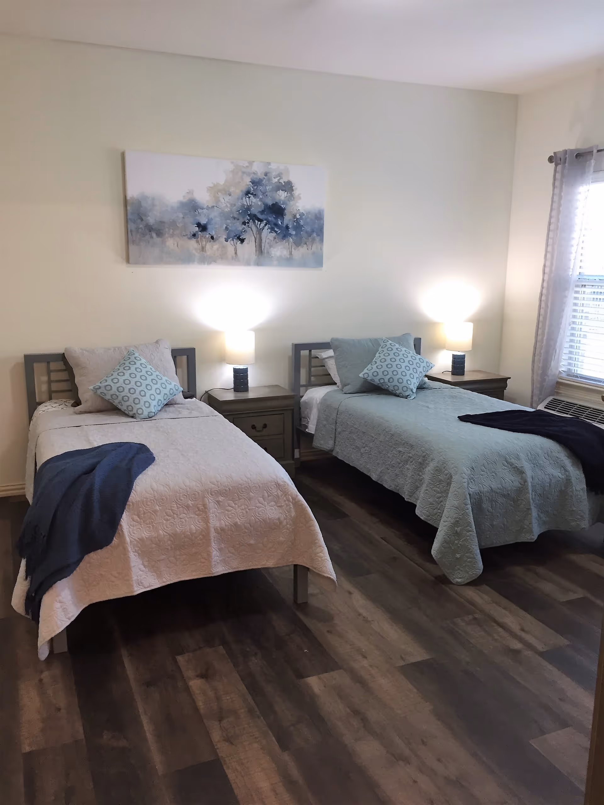 A bedroom with two twin beds, each with a pillow and a folded blanket at the foot. Between the beds is a nightstand with two lit lamps. A window with sheer curtains is on the right wall, and a painting of trees in blue tones hangs above the nightstand.