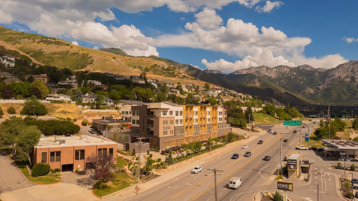 Aerial exterior view of a multi-story senior living building along a main road with homes and mountains in the background under a partly cloudy sky.