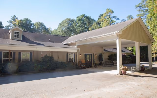 Exterior view of a single-story building with a covered entrance supported by white columns. The building has beige siding, a dark shingled roof, and several windows with dark shutters. There are rocking chairs and potted plants near the entrance. Trees and greenery are visible in the background under a clear blue sky.