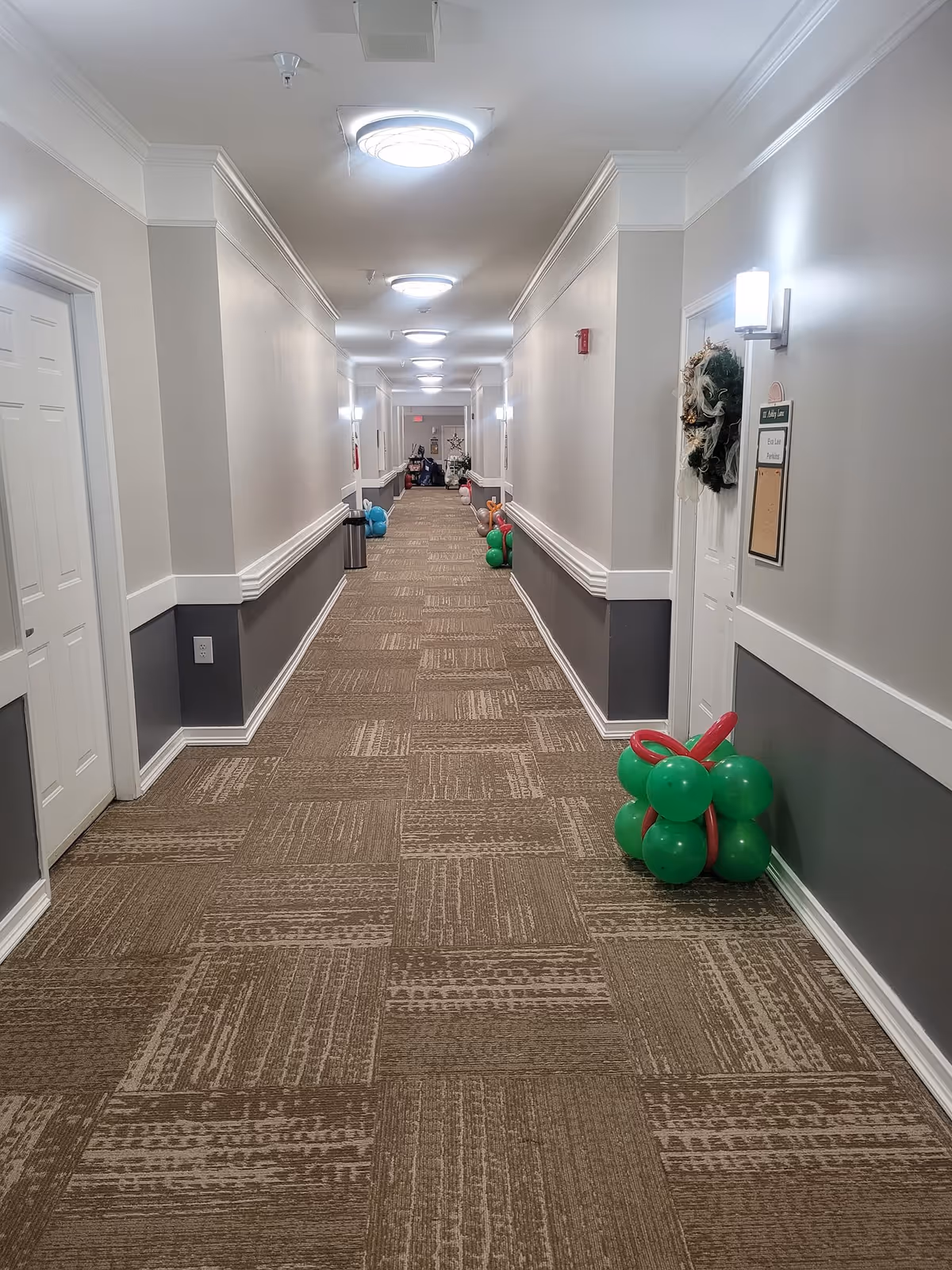 A long, well-lit hallway in a senior living facility with beige and gray walls, carpeted floor, and multiple closed doors on both sides. The hallway is decorated with clusters of colorful balloons and a wreath on one door.