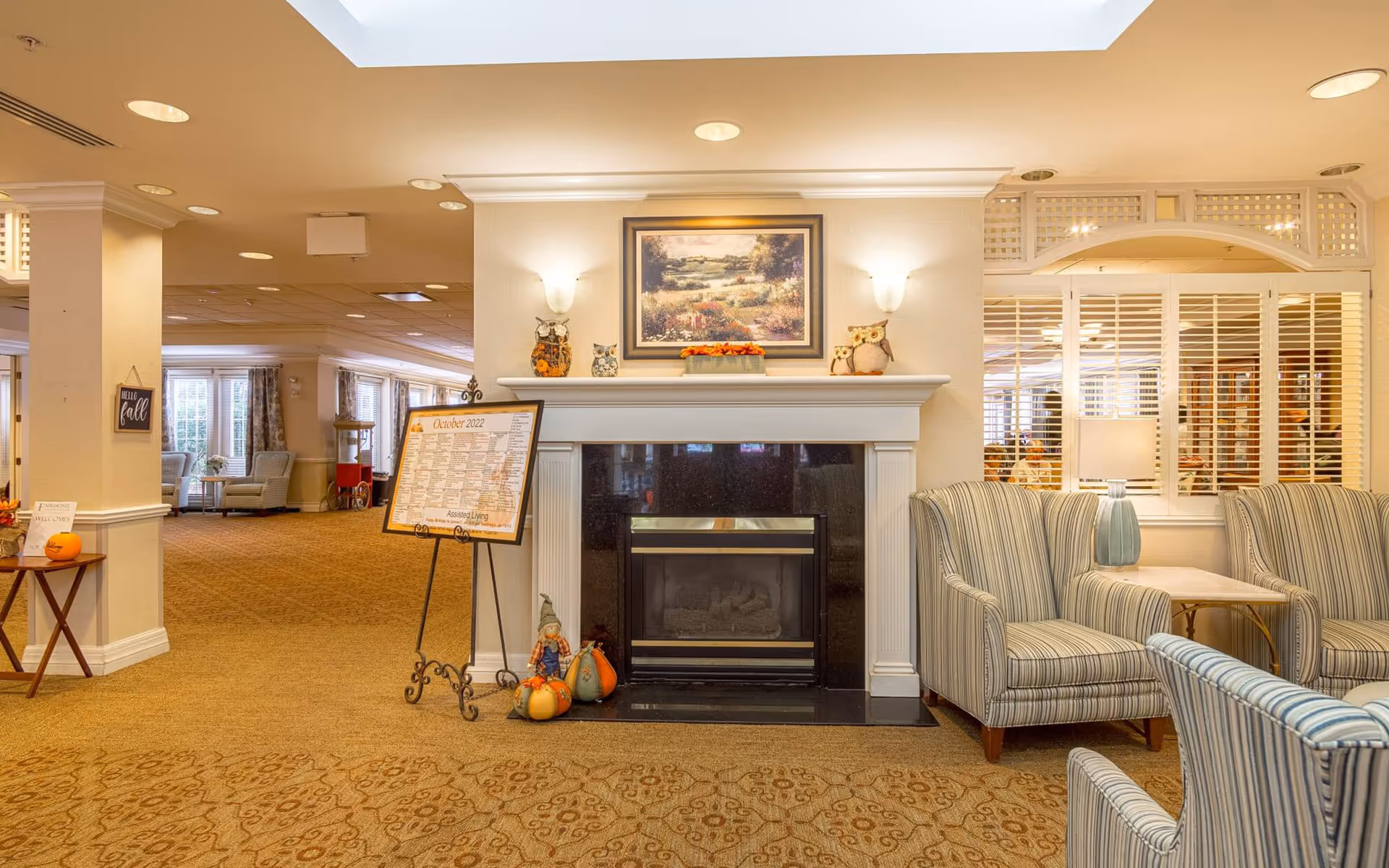 Lobby/lounge area of a senior living facility featuring a fireplace, striped armchairs, fall decorations, and an activities board.