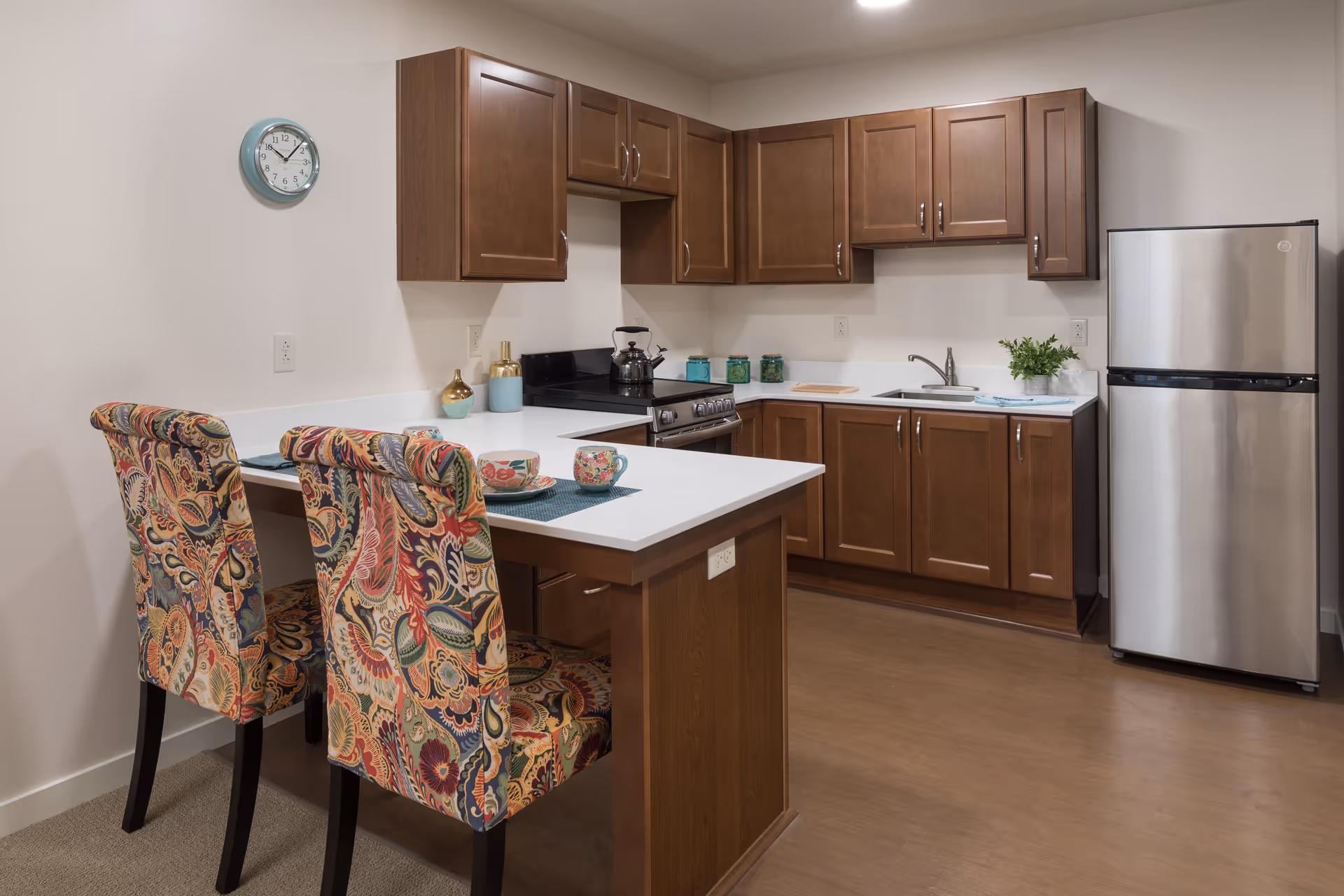 A modern kitchen with wooden cabinets, a stainless steel refrigerator, a stove with a kettle, and a white countertop island with two colorful patterned chairs. There are decorative items on the counter including a small plant and jars, and a wall clock is visible on the left wall.