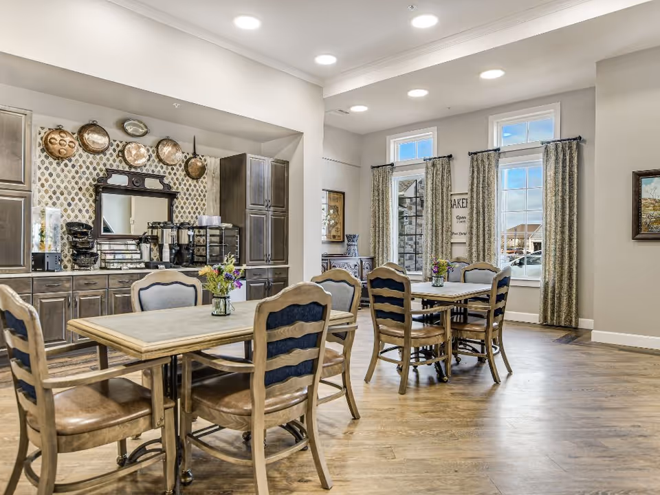 Bright communal dining room with wooden tables and chairs, a coffee station and cabinetry along one wall, and large windows with curtains.