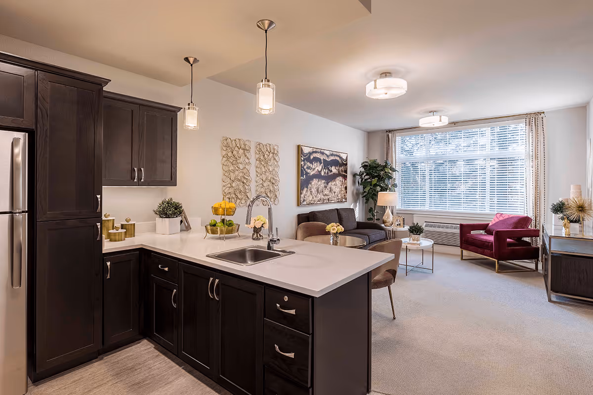 A modern open-concept living space featuring a kitchen with dark wood cabinets, a white countertop island with a sink, and stainless steel refrigerator. The living area includes a dark sofa, a red armchair, a coffee table, a side table with a lamp, and large windows with blinds allowing natural light to fill the room. Decorative wall art and plants add a cozy touch.