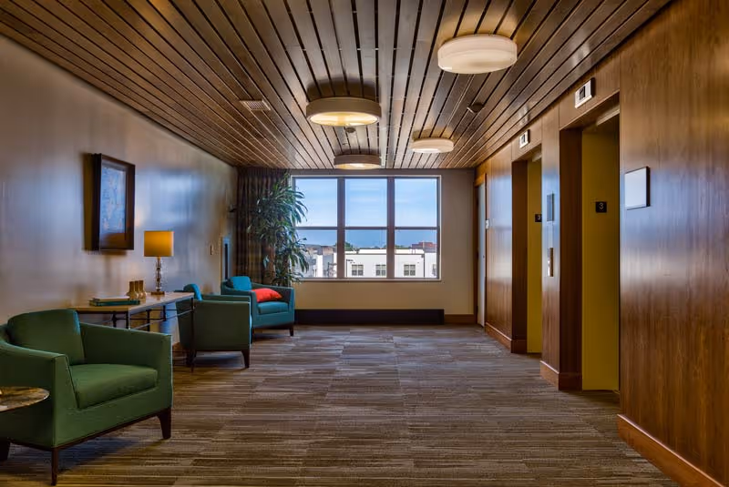 Carpeted senior living facility hallway with green lounge chairs and a table lamp, a large window at the end, and elevators along the right wall.