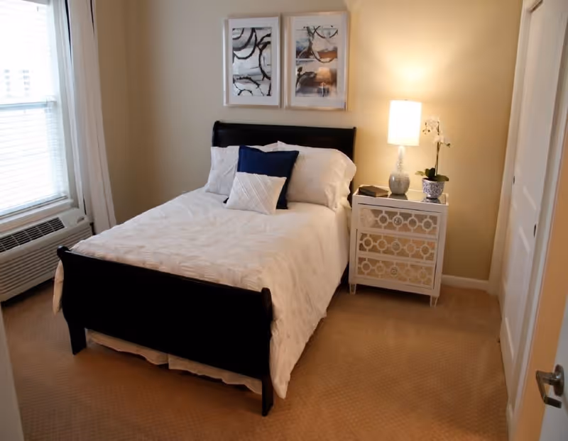 A neatly made bed with white bedding and two decorative pillows, one navy blue and one white, in a bedroom. There is a white nightstand with a decorative lamp, a small plant, and a book on it. Two abstract framed artworks hang on the beige wall above the bed. A window with white blinds and curtains is on the left side, and a door is partially visible on the right.