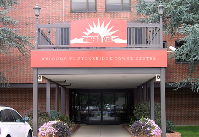 Entrance to a brick building with a covered walkway supported by columns. Above the entrance is a red sign with a white graphic of buildings and a sunburst, and text that reads 'WELCOME TO STONERIDGE TOWNE CENTRE'. There are two lampposts on either side of the entrance and flower beds with pink and purple flowers near the walkway.