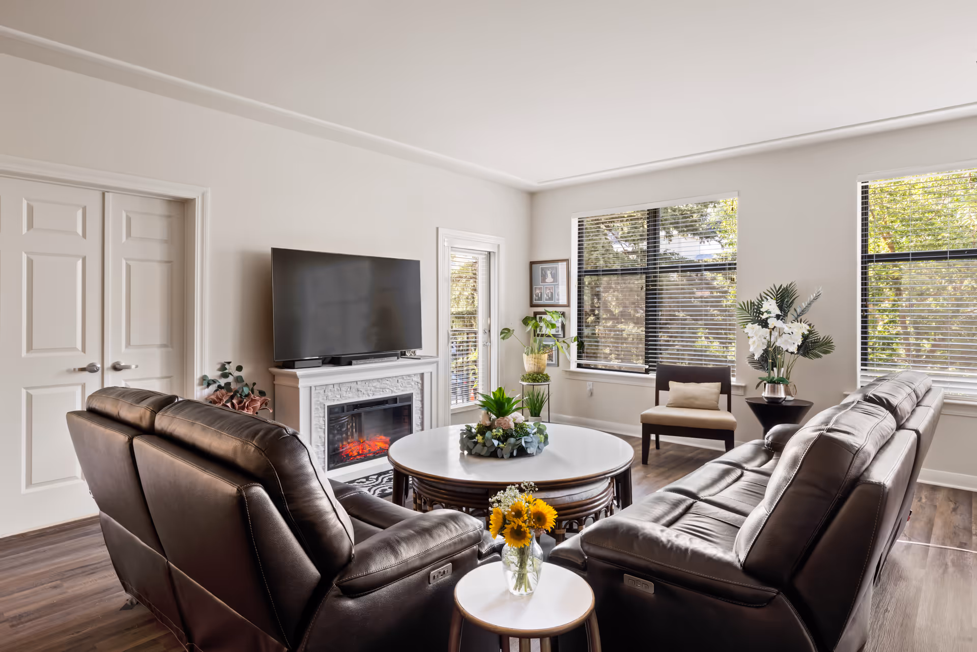 A bright living room with two dark brown leather sofas facing a white round coffee table decorated with plants. A flat-screen TV is mounted above a white electric fireplace. There are large windows with blinds letting in natural light, a small side table with a vase of sunflowers, and a single chair with a cushion near the windows.