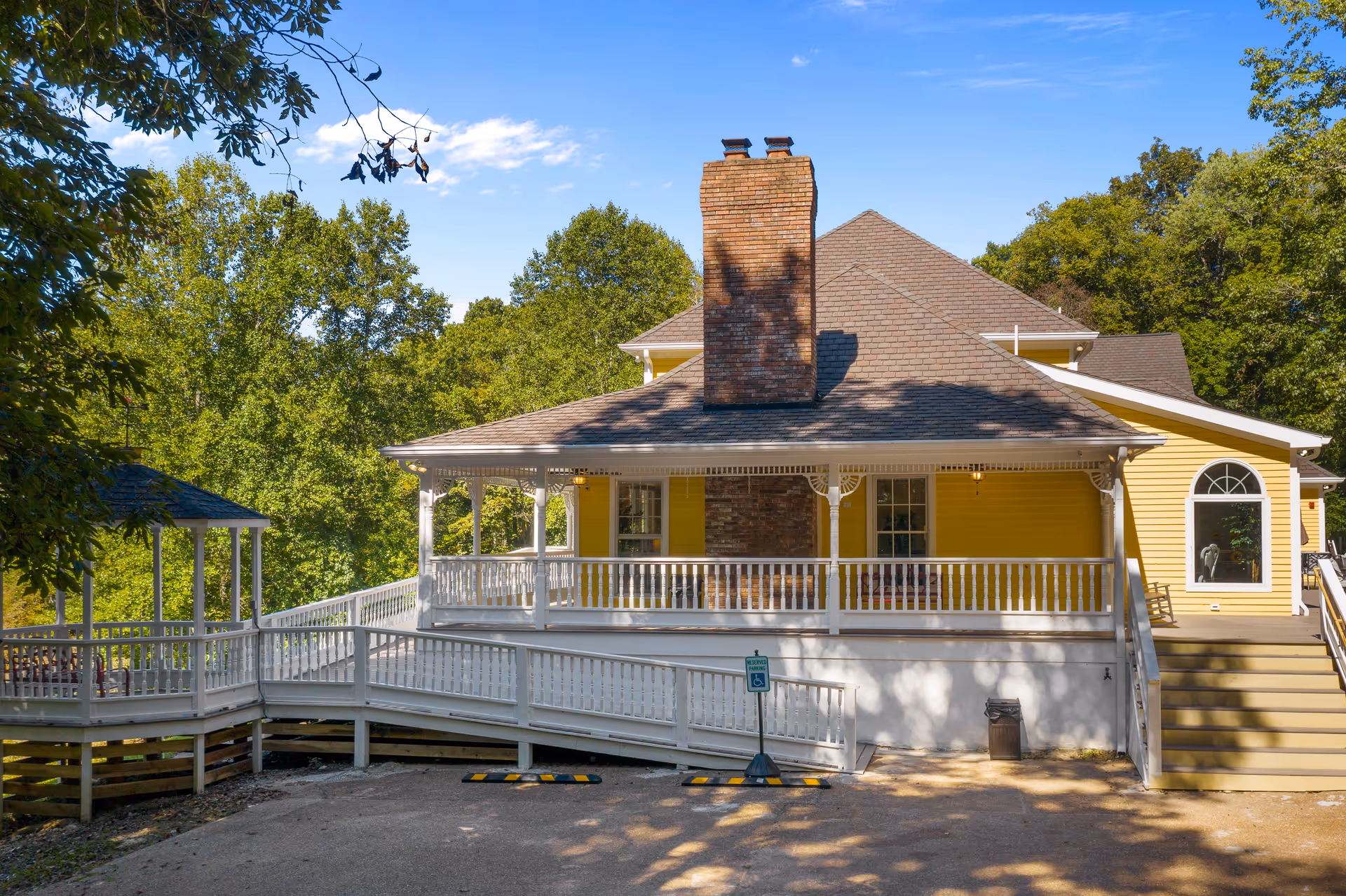Exterior view of a yellow senior living facility building with a large porch featuring white railings and a brick chimney. There is a wheelchair accessible ramp leading to the porch and a reserved parking sign for handicapped parking in front. The building is surrounded by green trees under a clear blue sky.
