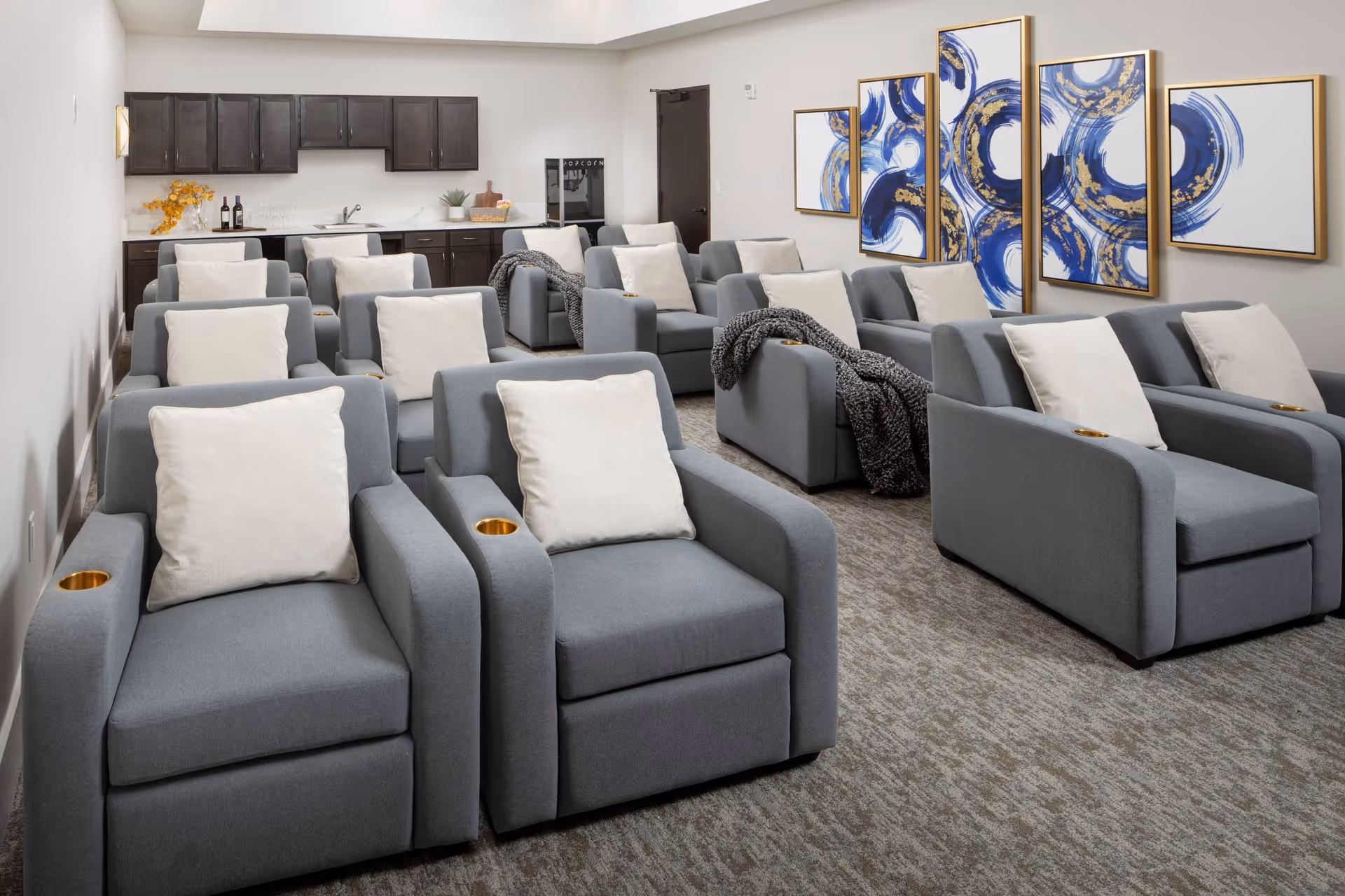 A cozy screening lounge with rows of gray reclining chairs with white pillows, a kitchenette at the back, and blue abstract art on the wall.