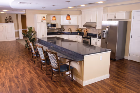 A spacious kitchen area with a large dark granite countertop island surrounded by four wooden chairs with patterned cushions. The kitchen features white cabinetry, stainless steel refrigerator, microwave, and stove. The floor is wood laminate, and there are three pendant lights hanging above the island. In the background, there is a plant and additional cabinetry.