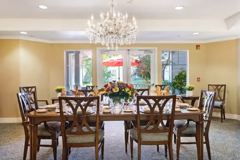A formal dining room with a large wooden table set for a meal, surrounded by eight wooden chairs with cushioned seats. A crystal chandelier hangs above the table, and a vase with colorful flowers is placed in the center. The room has beige walls, carpeted floor, and large glass doors and windows that show an outdoor area with greenery and a red umbrella.