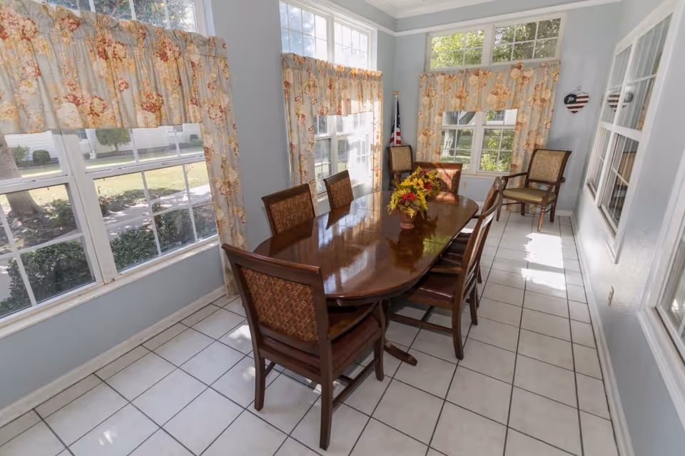 A bright dining room with large windows on two sides, covered with floral curtains. The room features a polished wooden dining table with six matching chairs, a vase with flowers on the table, and two additional chairs near the far wall. The floor is tiled in white, and an American flag is visible in the corner.