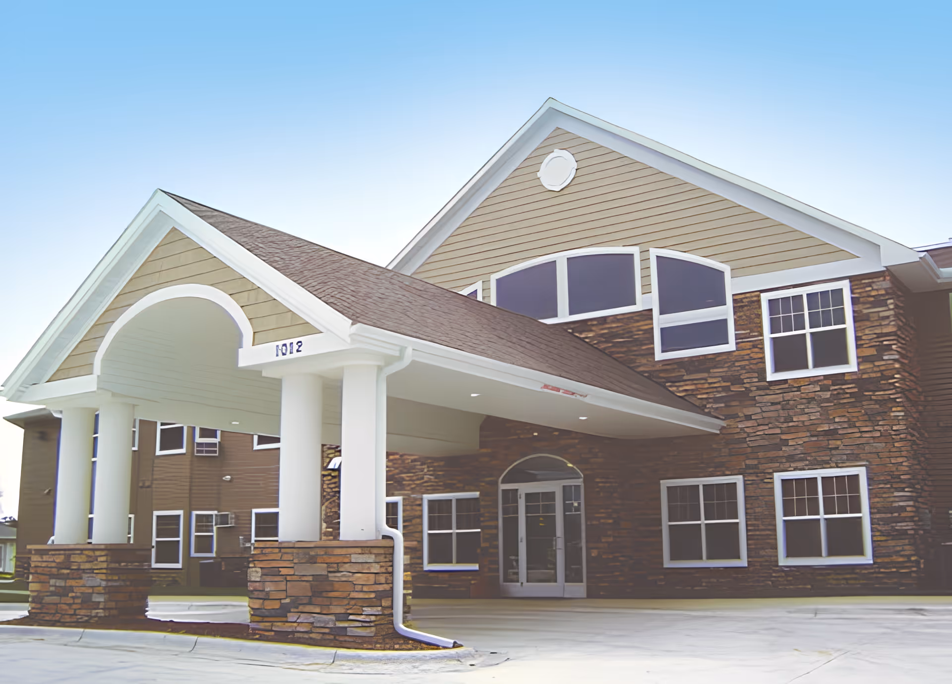 Exterior view of a senior living facility building with a covered entrance supported by white columns and stone bases, multiple windows, and a clear blue sky.