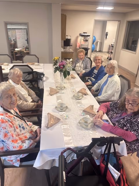 A group of six elderly women sitting around a long dining table set with cups, saucers, and folded napkins. The table has a white tablecloth and a vase with flowers in the center. The room appears to be a communal dining area in a senior living facility.