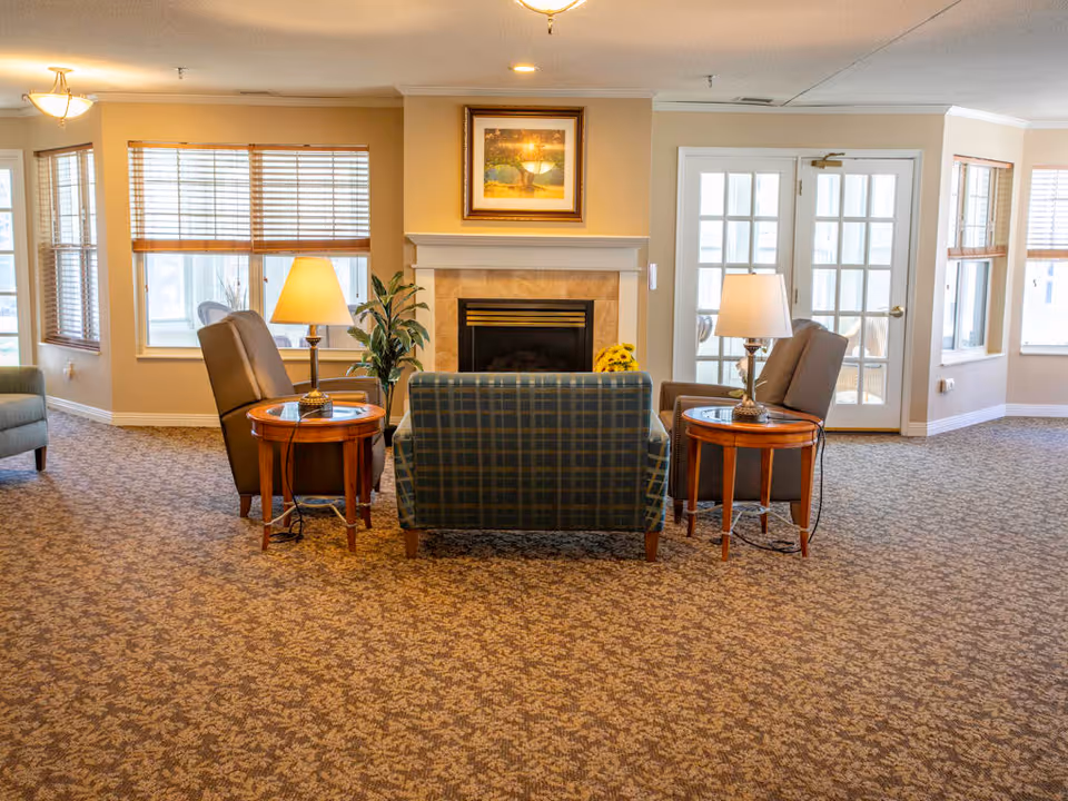 A cozy living room area with a patterned carpet, two armchairs, a plaid loveseat, and two wooden side tables with lamps. There is a fireplace with a framed picture above it, windows with wooden blinds, and a set of French doors leading to another room.