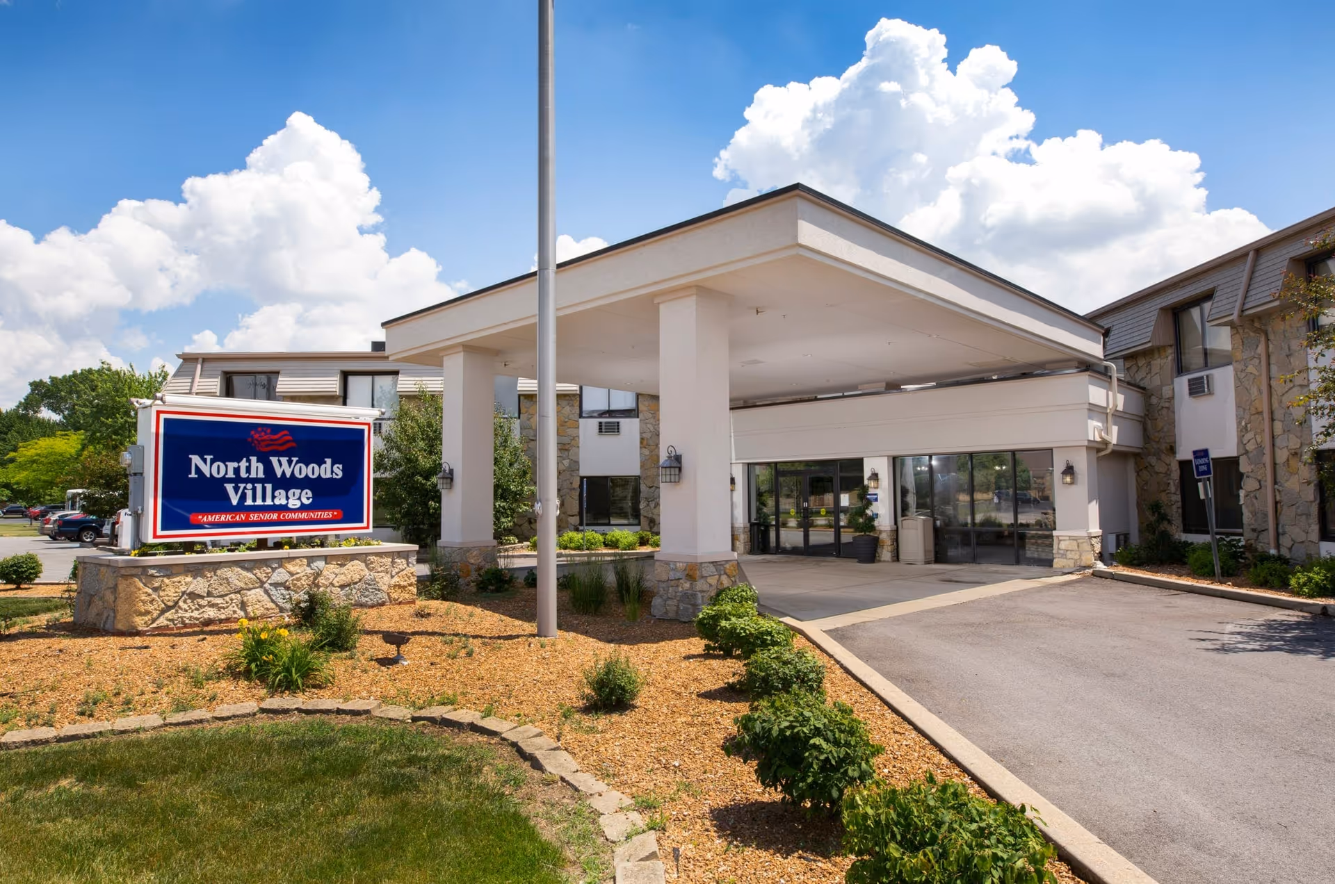 Front entrance of North Woods Village senior living facility with a covered drive-up canopy and a sign in the landscaped forecourt.