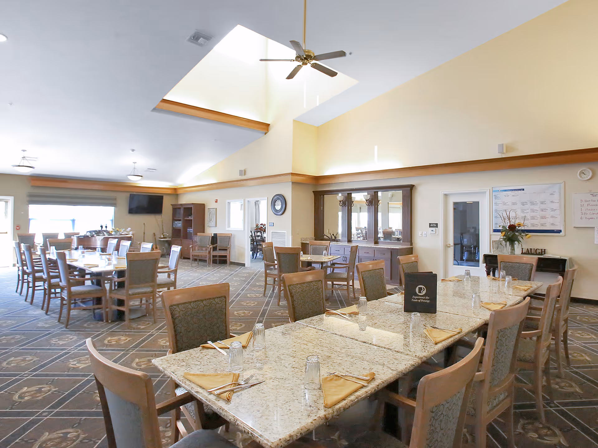 A spacious dining room in a senior living facility with multiple tables and chairs arranged neatly. The tables are set with glasses, napkins, and silverware. The room has high ceilings with ceiling fans, large windows allowing natural light, and a carpeted floor with a patterned design. There is a cabinet with a mirror and some decorations on the wall.