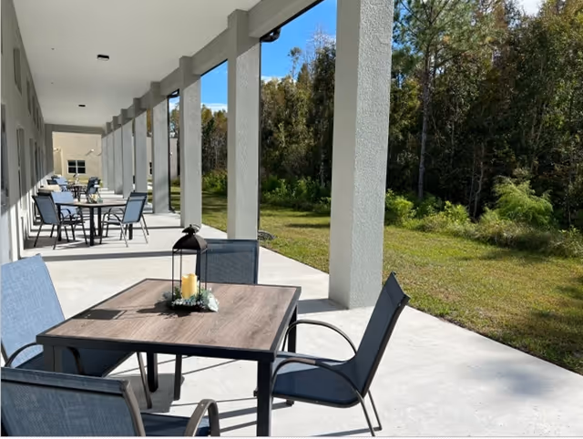 Covered outdoor patio with dining tables and chairs facing a grassy yard and trees.