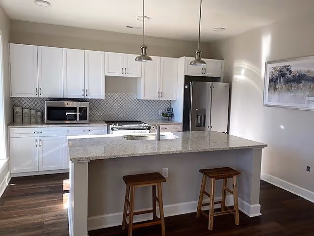 A modern kitchen with white cabinets, a granite countertop island with two wooden stools, stainless steel refrigerator, microwave, and stove. The kitchen has pendant lights hanging above the island and a framed artwork on the wall to the right.