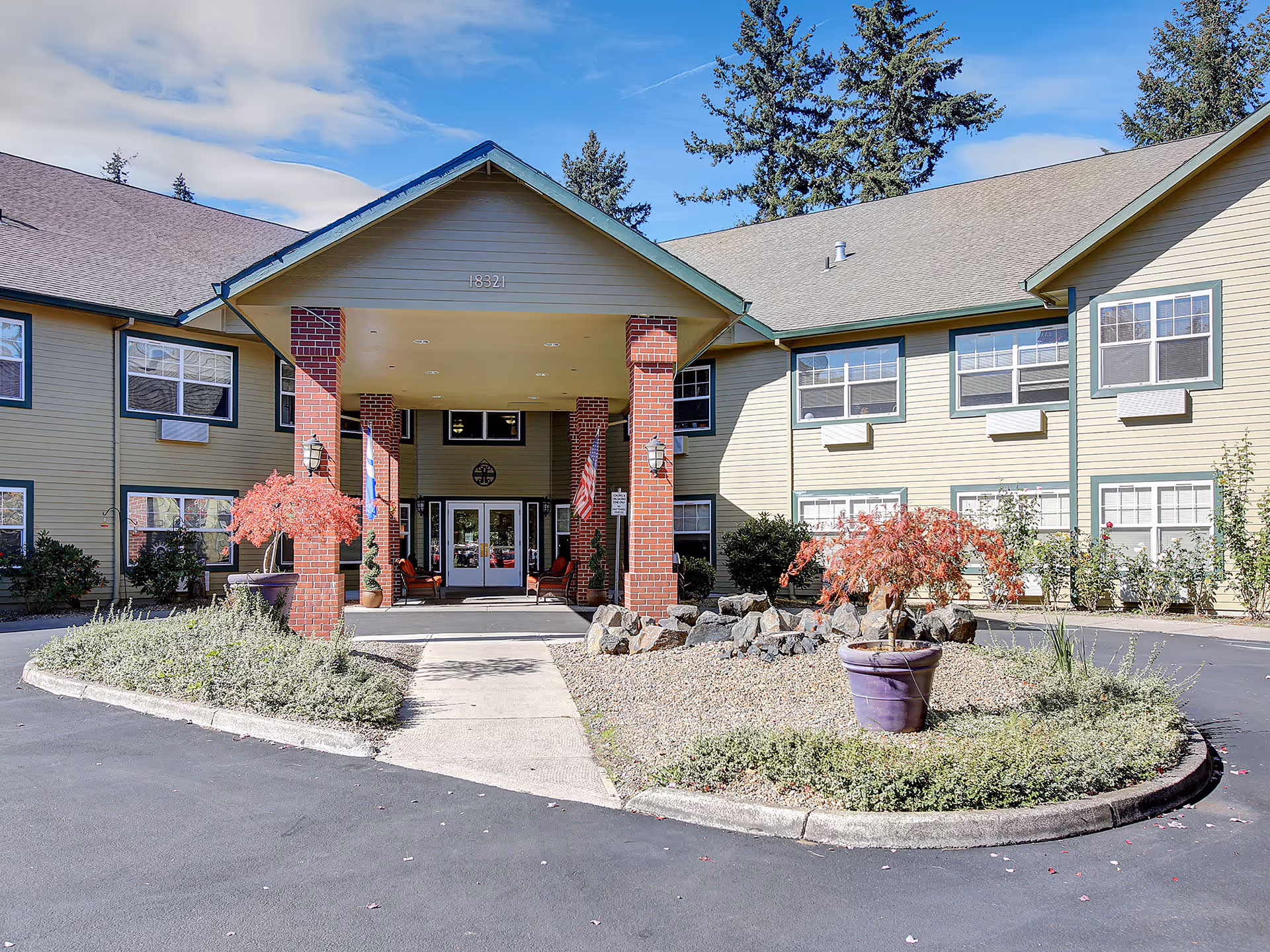 Front exterior view of Prestige Senior Living Riverwood building with a covered entrance supported by brick pillars, surrounded by landscaped plants and trees under a partly cloudy sky.