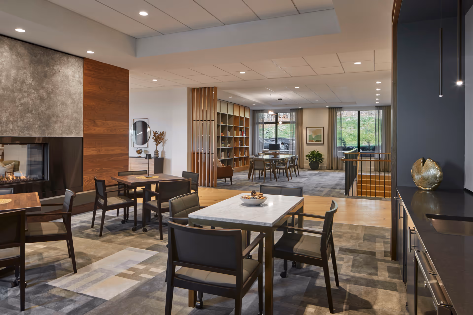 A modern senior living facility common area with multiple tables and chairs arranged for seating. The room features a fireplace with a textured wall, a bookshelf with decorative items, large windows with curtains letting in natural light, and a small kitchenette area with a sink and countertop. The space has a warm and inviting atmosphere with neutral tones and contemporary furnishings.