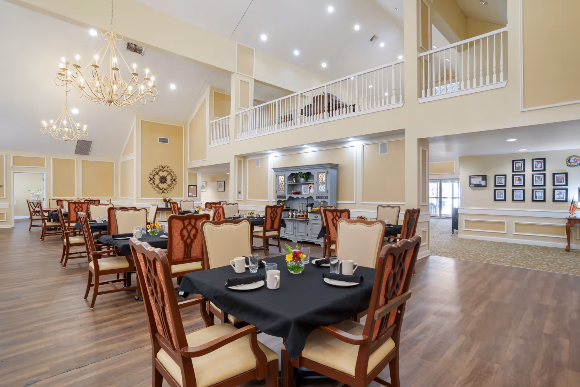 A spacious dining room with multiple tables covered in black tablecloths, each set with white cups, plates, and silverware. The room features elegant wooden chairs with cream and reddish upholstery, chandeliers hanging from a high ceiling, and a light beige color scheme with white trim. A blue hutch with decorative items is against the far wall, and a hallway with framed pictures is visible in the background.