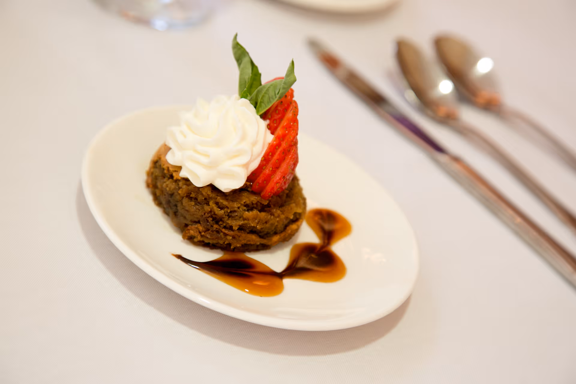 A small dessert on a white plate featuring a brown cake topped with whipped cream, a sliced strawberry, and a green leaf garnish, with decorative chocolate sauce on the plate. In the background, there are three pieces of silver cutlery on a white tablecloth.
