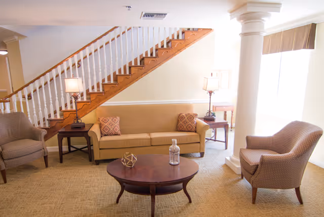 A cozy living room area with a beige sofa, two patterned armchairs, a round wooden coffee table with decorative items, two side tables with lamps, a staircase with wooden steps and white railing, and a large window with blinds letting in natural light.