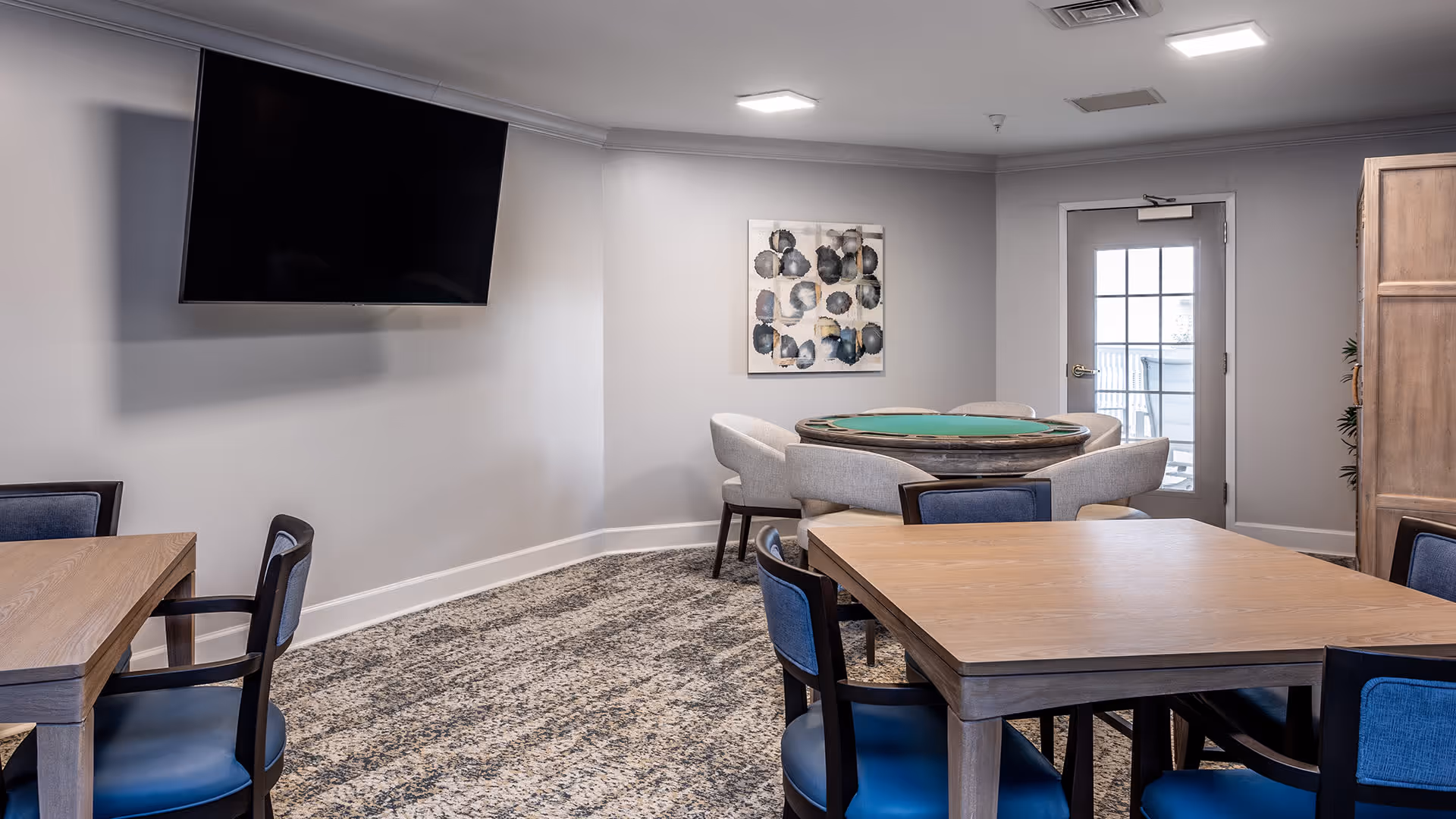 A well-lit common room with multiple wooden tables and blue cushioned chairs. A round table with a green felt top and four gray upholstered chairs is positioned near a door with glass panes. A large flat-screen TV is mounted on the wall, and abstract artwork hangs on the far wall. The room has a patterned carpet and neutral-colored walls.