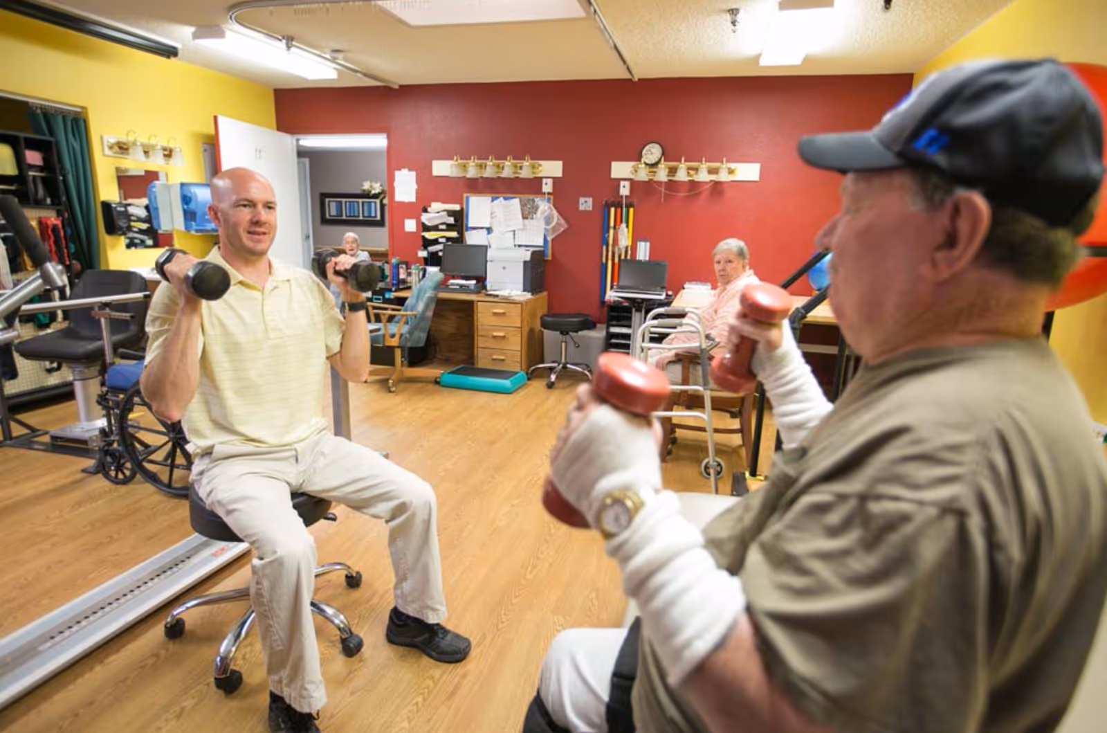 Two men lifting dumbbells while seated in a rehabilitation or exercise room with wooden floors and yellow and red walls. In the background, two elderly women are seated, one with a walker. The room contains office desks, chairs, and exercise equipment.