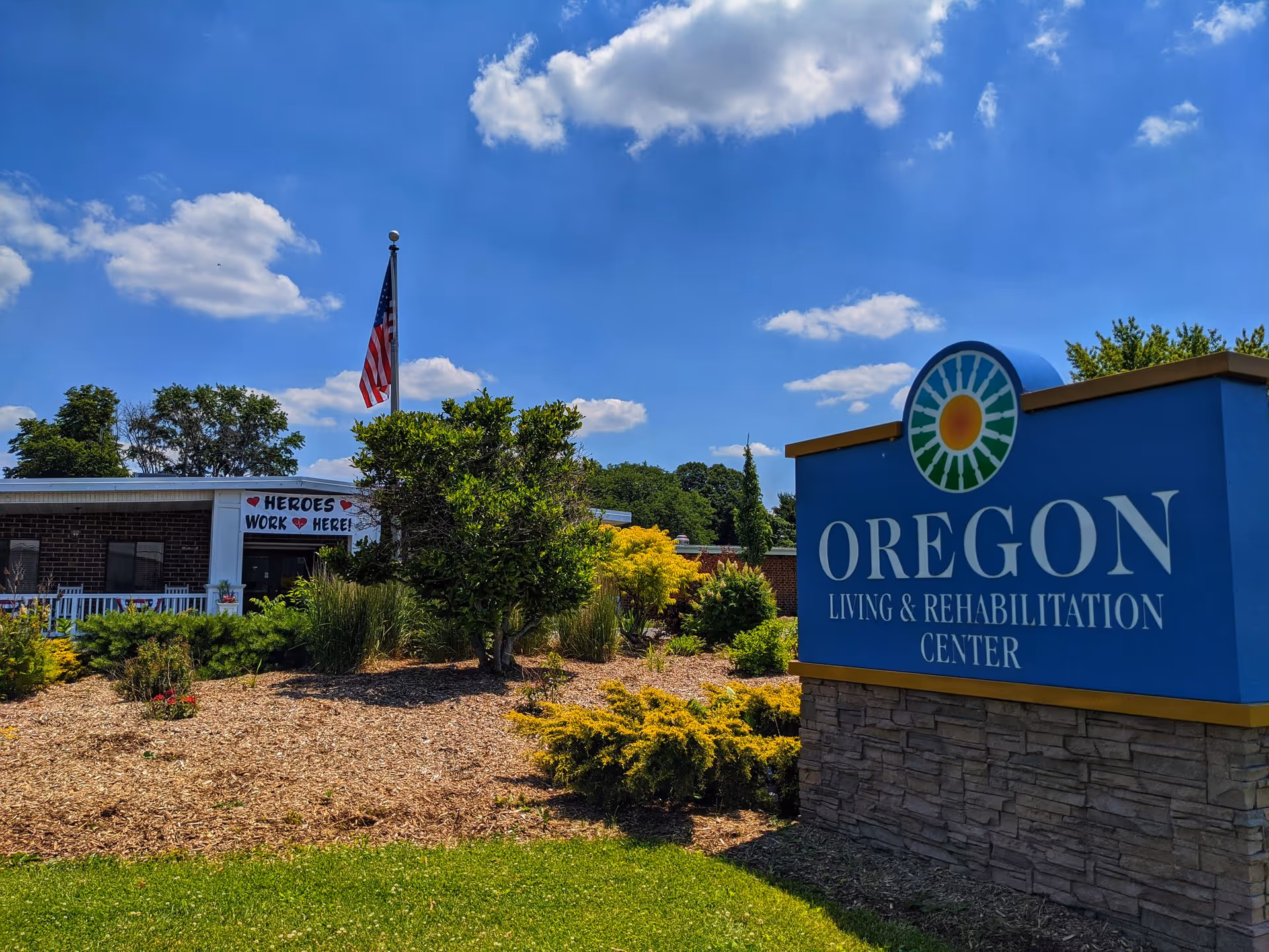 Exterior view of Oregon Living & Rehabilitation Center on a sunny day with a blue sky and scattered clouds. The building is partially visible behind landscaped bushes and trees. An American flag is flying on a flagpole near the entrance, where a sign reads 'HEROES WORK HERE!'. A large blue sign with the facility's name and logo is prominently displayed in the foreground.