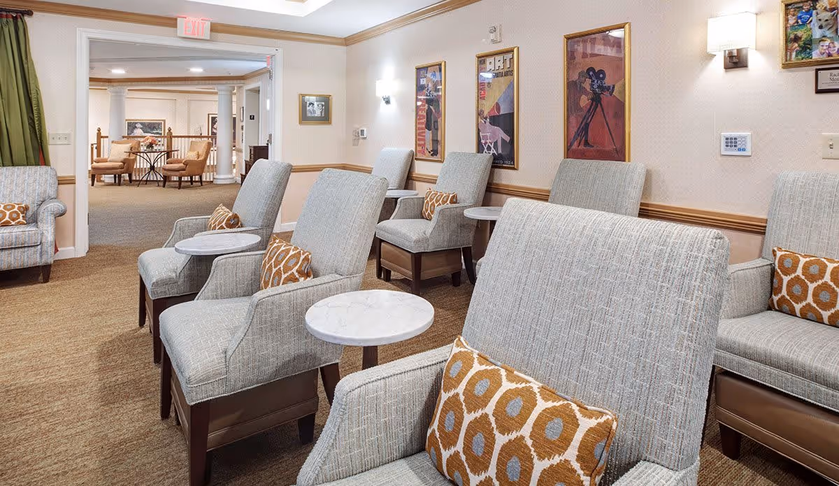 A cozy senior living common area with several comfortable armchairs arranged in rows, each with a small round side table and patterned cushions. The walls are decorated with framed vintage-style movie posters and a few small wall lamps. In the background, there is an open doorway leading to another seating area with more chairs and a table.