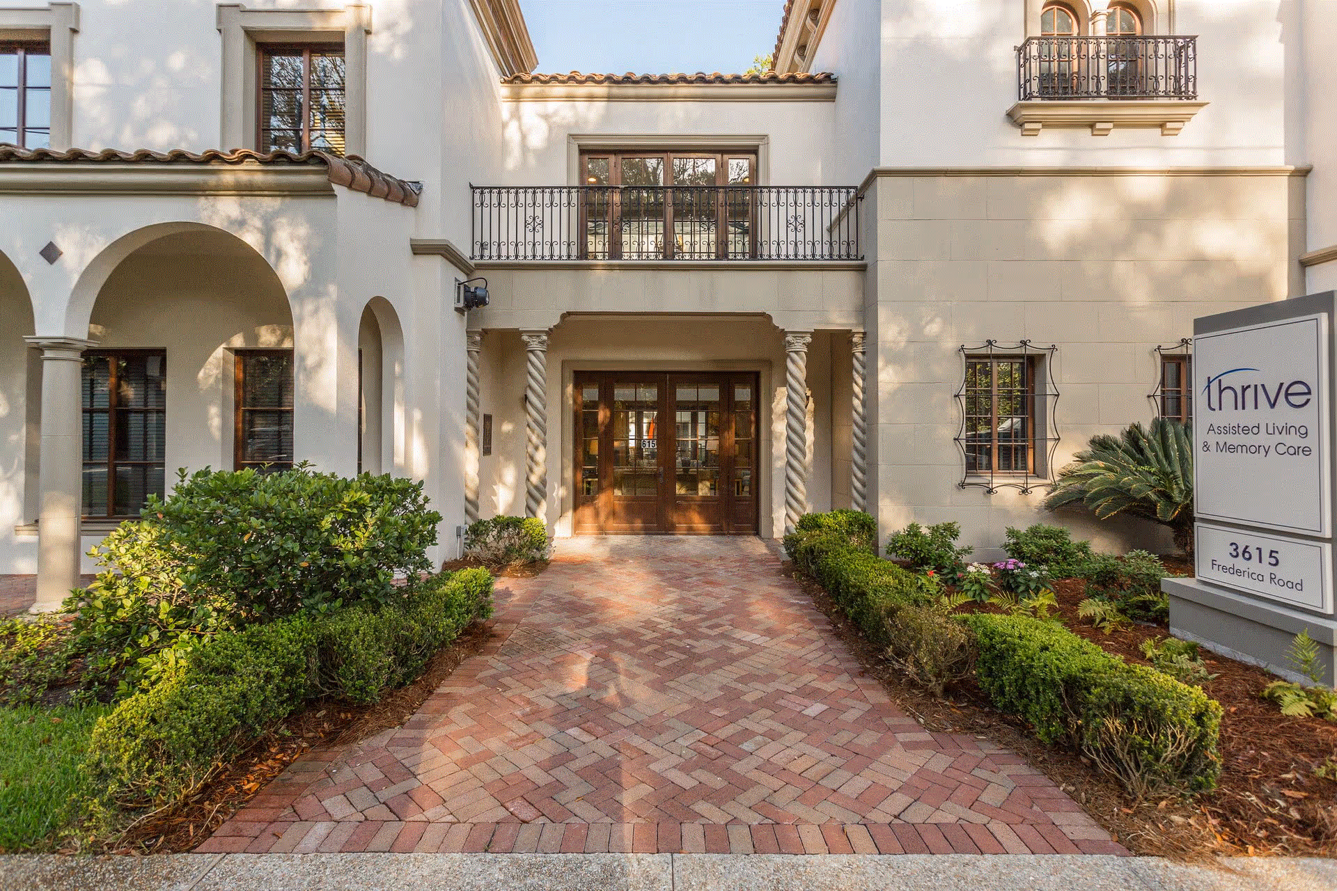 Front entrance of a senior living facility with a brick walkway leading to double wooden doors. The building has a beige exterior with decorative columns and balconies. There are neatly trimmed bushes and plants along the walkway. A sign on the right reads 'thrive Assisted Living & Memory Care 3615 Frederica Road'.