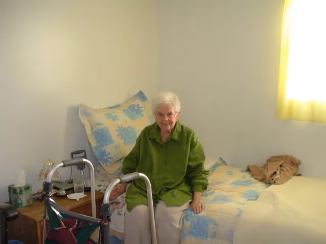 An elderly woman with white hair and glasses sitting on the edge of a bed in a bedroom. The bed has a floral patterned bedspread in blue and beige tones. Next to the bed is a wooden nightstand with a box of tissues, a glass of water, and some personal items. A walker is positioned in front of the woman. The room has plain light-colored walls and a window with yellow curtains letting in natural light.